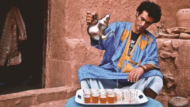A Moroccan man dressed in a traditional blue Sahrawi clothing with yellow patterns is pouring Moroccan tea from a height