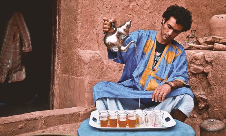 A Moroccan man dressed in a traditional blue Sahrawi clothing with yellow patterns is pouring Moroccan tea from a height